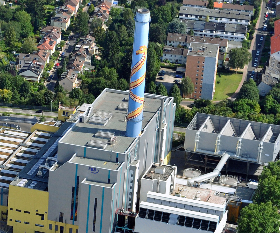 Luftaufnahme einer Industrieanlage mit hohem Schornstein und dem FES-Logo, umgeben von Wohngebäuden.